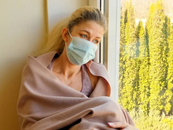 医療マスクのブロンドの女の子は悲しいです 毛布で覆われ 窓の外を見て ストック画像