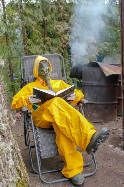 Gaz maskeli ve sarı koruyucu kıyafetli bir insan oturur ve kitap okur.