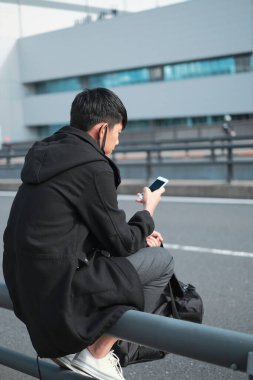 Yol bariyerinde oturan Asyalı genç adam akıllı telefon kullanırken otobüs ve taksi şoförü konseptini bekliyor.