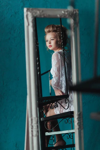 Spiegelung im Spiegel bescheidene schöne blonde Braut in weißem Negligee sitzt auf der schwarzen schmiedeeisernen Treppe. — Stockfoto