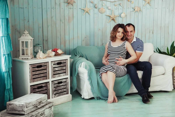 Giovane donna incinta con marito su divano bianco in camera blu. coppia vestita con abiti a righe. Estate ispirazione vacanza composizione sfondo — Foto Stock