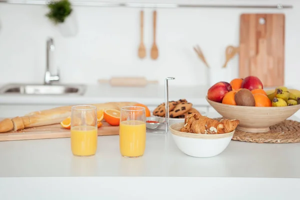 Placa de sabrosas galletas con migas de chocolate, croissant, bolsita, plátanos, manzanas, caqui, naranja y zumo de naranja sobre mesa blanca — Foto de Stock