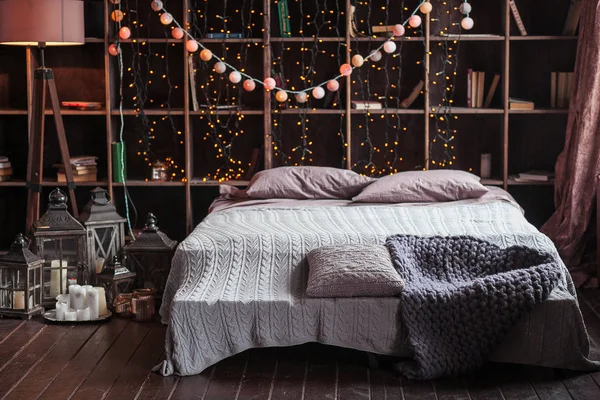 coziness, comfort, interior and holidays concept - cozy bedroom with bed and garland lights at home. A rack with books behind the bed. Candles, a lamp and a lamp stand near the bed. Plaid hand-knitted