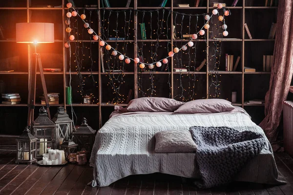 Aconchego, conforto, interior e férias conceito quarto acolhedor com cama e luzes guirlanda em casa. Uma prateleira com livros atrás da cama. Velas, uma lâmpada e um suporte de lâmpada perto da cama. Xadrez de malha manual — Fotografia de Stock