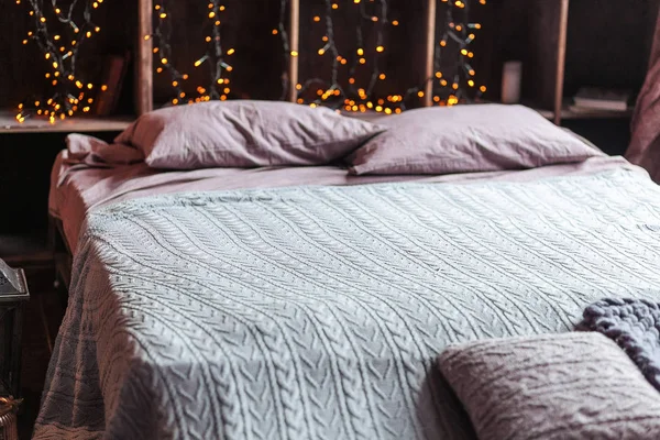 Coziness, comfort, interior and holidays concept - cozy bedroom with bed and garland lights at home. A rack with books behind the bed. Candles, a lamp and a lamp stand near the bed. Plaid hand-knitted — Stock Photo, Image
