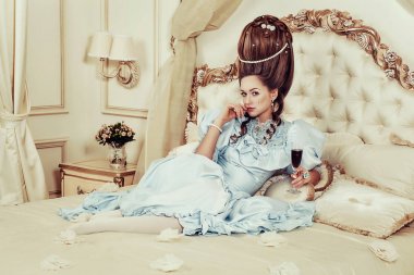 Indoors shot in the Marie Antoinette style. A young sexy girl in a lush blue retro dress with a high hairstyle lies on the bed with a glass of sparkling wine in her hand. Woman tired of luxury clipart