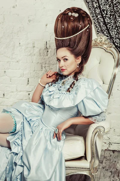 Tiro interior al estilo María Antonieta. Una chica sexy joven en un exuberante vestido retro azul con un peinado alto se sienta en el sofá. Mujer cansada del lujo — Foto de Stock