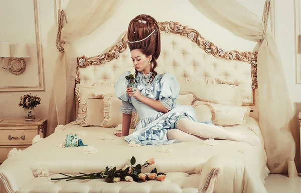 Tiro interior al estilo María Antonieta. Una chica sexy joven en un exuberante vestido retro azul con un peinado alto se encuentra en la cama con una flor en la mano. Mujer cansada del lujo — Foto de Stock