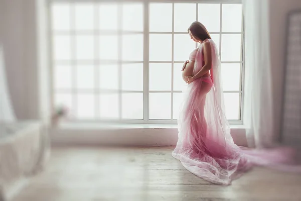 Jovem grávida em tecido rosa está em pé junto à janela. Mulher grávida irreconhecível estúdio tiro — Fotografia de Stock