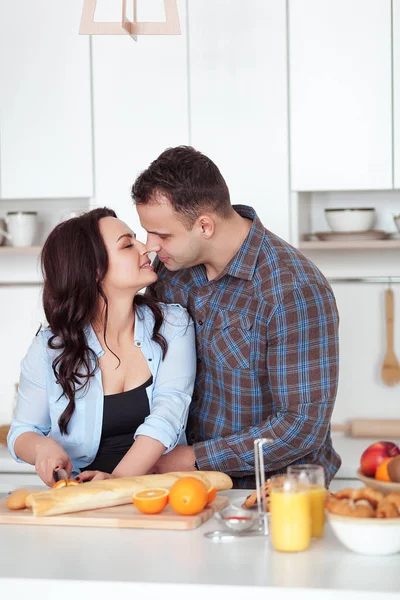 Пара, готовящая свежий органический сок на кухне вместе. Молодая женщина в голубой рубашке режет багет. Мужчина обнимает свою девушку на белой кухне. Романтическая концепция — стоковое фото