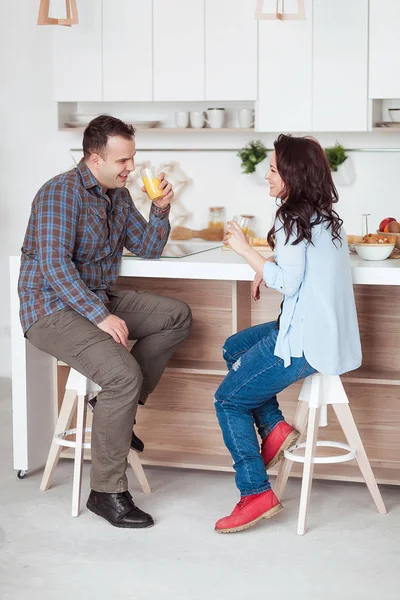 Atractiva pareja sentada tomando jugo juntos y riendo en casa en la cocina — Foto de Stock