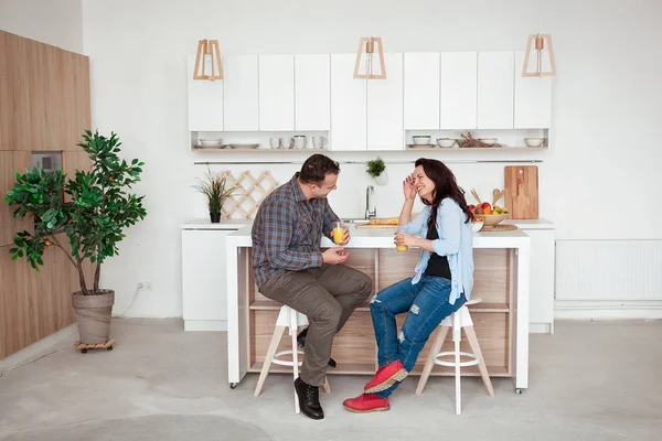 Atractiva pareja sentada tomando jugo juntos y riendo en casa en la cocina —  Fotos de Stock