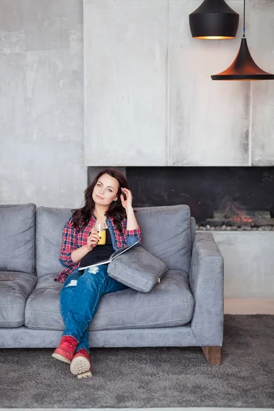 Mujer joven relajándose en casa con un vaso de jugo de naranja. Hermosa chica en un estilo casual se sienta en el sofá y lee un libro en la casa de estilo loft — Foto de Stock