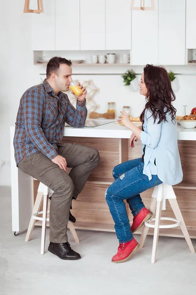 Atractiva pareja sentada tomando jugo juntos y riendo en casa en la cocina —  Fotos de Stock