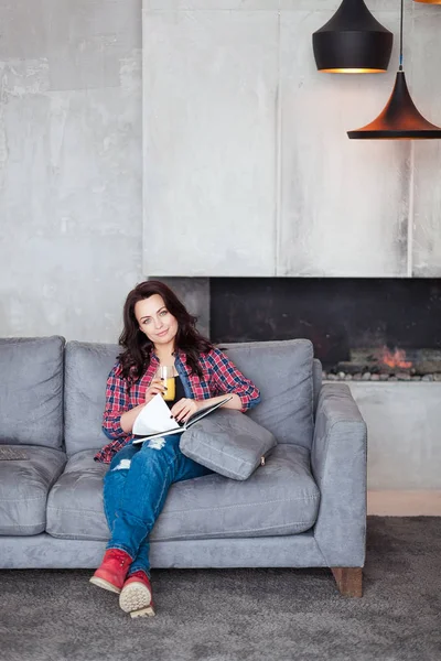 Mujer joven relajándose en casa con un vaso de jugo de naranja. Hermosa chica en un estilo casual se sienta en el sofá y lee un libro en la casa de estilo loft — Foto de Stock