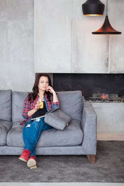 Mujer joven relajándose en casa con un vaso de jugo de naranja. Hermosa chica en un estilo casual se sienta en el sofá y lee un libro en la casa de estilo loft — Foto de Stock