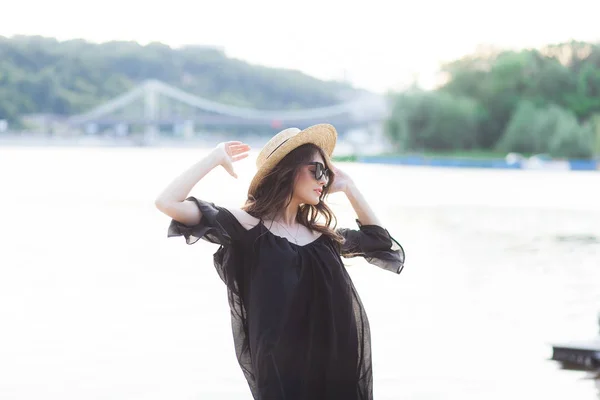 Mooie jonge brunette vrouw in zwarte jurk, zwarte zonnebrillen en stro hoed lopen op het zand op het strand en poseren bij zonsopgang in de zomer — Stockfoto