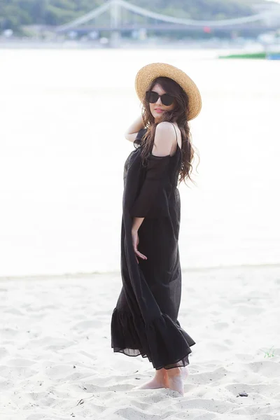 Linda jovem morena de vestido preto, óculos de sol pretos e chapéu de palha andando na areia na praia e posando ao nascer do sol no verão — Fotografia de Stock