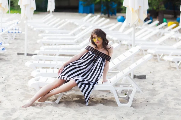 Retrato de jovem morena feliz em preto e branco vestido listrado sentado no leito solar enquanto relaxa na praia — Fotografia de Stock
