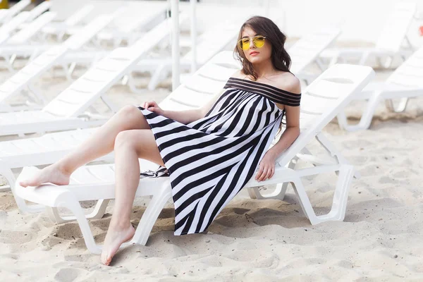 Portrait of young brunette woman in black and white striped dress and yellow sunglasses sitting on sunbed while relaxing on the beach — Stock Photo, Image