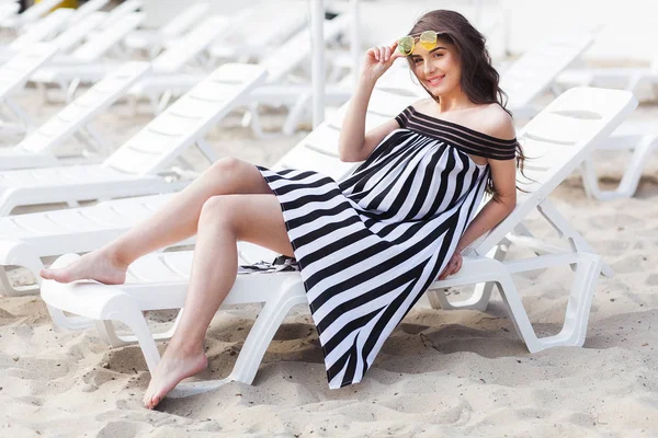 Portrait of happy young brunette woman in Black and white striped dress sitting on sunbed while relaxing on the beach — Stock Photo, Image