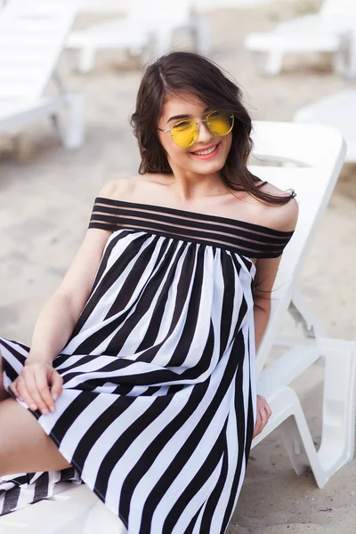 Retrato de jovem morena feliz em preto e branco vestido listrado sentado no leito solar enquanto relaxa na praia — Fotografia de Stock