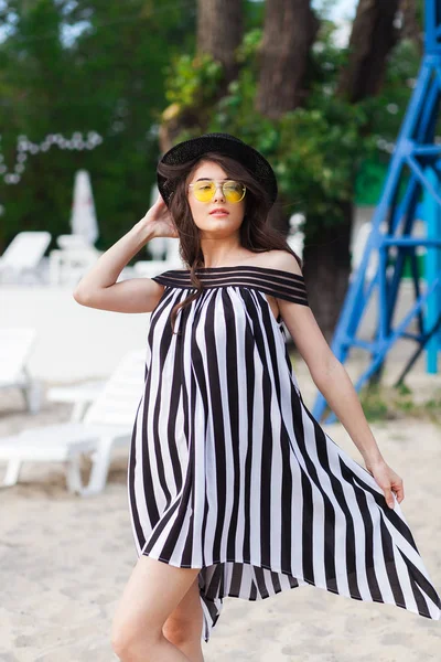 Mulher de viagem de luxo em roupas de praia preto e branco andando dando um passeio na praia de verão de areia. Menina turista em férias de verão segurando chapéu de sol e óculos de sol amarelos na estância de férias — Fotografia de Stock