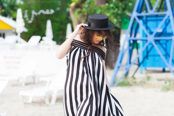 Mulher de viagem de luxo em roupas de praia preto e branco andando dando um passeio na praia de verão de areia. Menina turista em férias de verão segurando chapéu de sol e óculos de sol amarelos na estância de férias — Fotografia de Stock