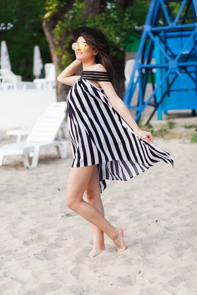 Mulher de viagem de luxo em roupas de praia preto e branco andando dando um passeio na praia de verão de areia. Menina turista em férias de verão segurando chapéu de sol e óculos de sol amarelos na estância de férias — Fotografia de Stock