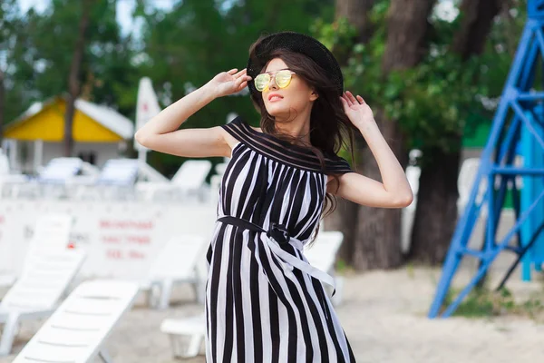 Femme de voyage de luxe en noir et blanc vêtements de plage marchant en se promenant sur la plage de sable d'été. Fille touristique en vacances d'été tenant chapeau de soleil et lunettes de soleil jaunes à la station de vacances — Photo