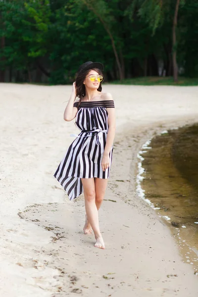 Mulher de viagem de luxo em roupas de praia preto e branco andando dando um passeio na praia de verão de areia. Menina turista em férias de verão segurando chapéu de sol e óculos de sol amarelos na estância de férias — Fotografia de Stock