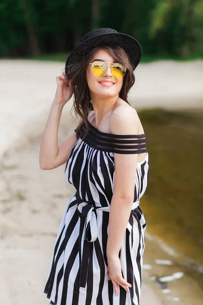 Luxury travel woman in black and white beachwear walking taking a stroll on sand summer beach. Girl tourist on summer holiday holding sun hat and yellow sunglasses at vacation resort — Stock Photo, Image