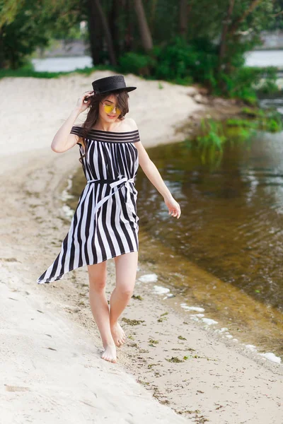 Mulher de viagem de luxo em roupas de praia preto e branco andando dando um passeio na praia de verão de areia. Menina turista em férias de verão segurando chapéu de sol e óculos de sol amarelos na estância de férias — Fotografia de Stock