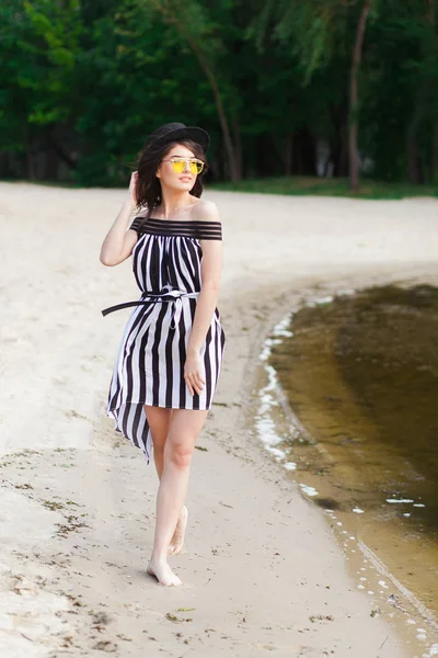 Mulher de viagem de luxo em roupas de praia preto e branco andando dando um passeio na praia de verão de areia. Menina turista em férias de verão segurando chapéu de sol e óculos de sol amarelos na estância de férias — Fotografia de Stock