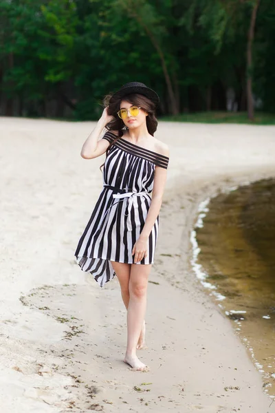 Mulher de viagem de luxo em roupas de praia preto e branco andando dando um passeio na praia de verão de areia. Menina turista em férias de verão segurando chapéu de sol e óculos de sol amarelos na estância de férias — Fotografia de Stock