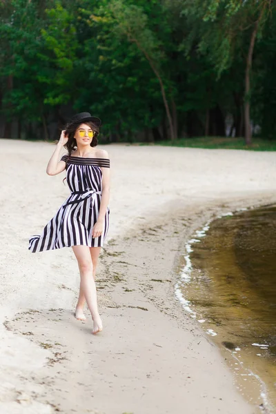 Luxusní cestovní žena v černé a bílé plavky pěší procházku na pláži písečné léto. Dívka turista na letní pobyty v rekreačním středisku drží slunce klobouk a žluté brýle — Stock fotografie
