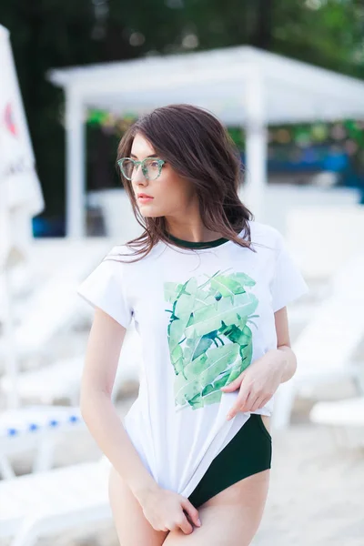 Retrato de moda de verano de mujer impresionante con cuerpo sexy en forma bronceada, con camiseta blanca con estampado verde, traje de baño y gafas de sol verdes con estilo, posando en la playa con arena — Foto de Stock