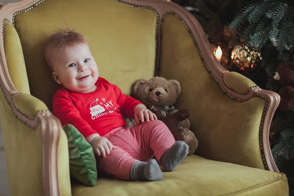 Menino bonito no sorriso pulôver vermelho enquanto sentado na poltrona antiquada perto da árvore de Natal no quarto. Fechar tiro — Fotografia de Stock
