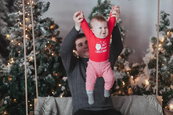 Pai e 1 ano de idade filho brincando na frente da árvore de Natal sentado no balanço de madeira no estúdio . — Fotografia de Stock