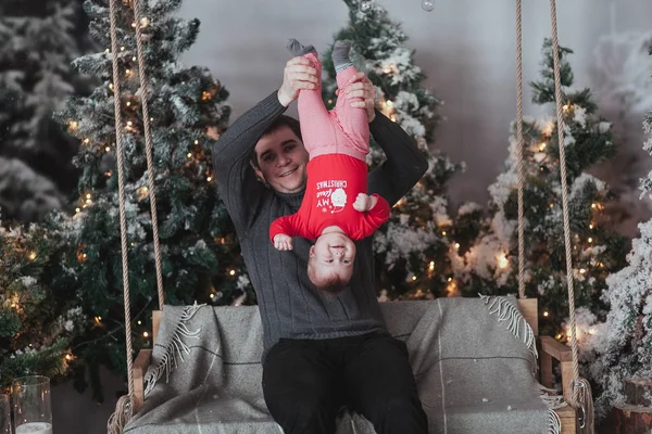 Pai e filho de 1 ano brincando na frente da árvore de Natal sentado no balanço de madeira no estúdio. Pai segurar criança virou de cabeça para baixo — Fotografia de Stock