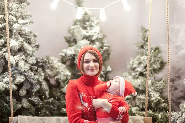 Mãe de família feliz e menino em santa chapéu perto de neve coberto árvores de Natal com sentado no balanço. guirlanda brilhante acima das cabeças — Fotografia de Stock