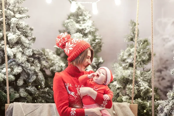 Mãe de família feliz e menino em santa chapéu perto de neve coberto árvores de Natal com sentado no balanço. guirlanda brilhante acima das cabeças — Fotografia de Stock