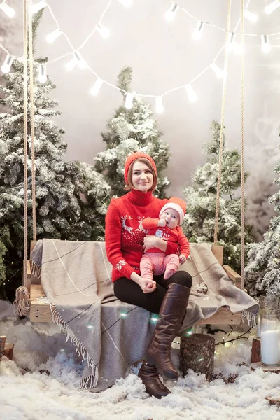 Conceito de Natal mãe e filho se senta em um balanço de madeira em pulôveres vermelhos e chapéus na frente de árvores cobertas de neve . — Fotografia de Stock