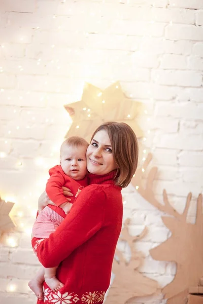 Uma jovem mulher segura bebê vestido para o evento de Natal posando parede de tijolo branco no fundo com luz guirlanda e decoração de veados de madeira — Fotografia de Stock