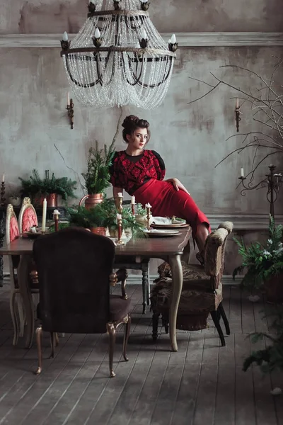 Mujer en vestido rojo vintage se sienta en la mesa servida para la cena de Navidad. Loft interior con pared de hormigón transparente — Foto de Stock
