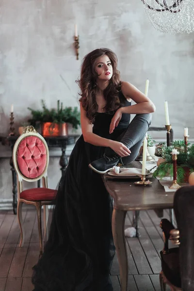 Joven hermosa dama en vestido de noche en el interior del loft se sienta en la mesa con velas y ramas de abeto — Foto de Stock