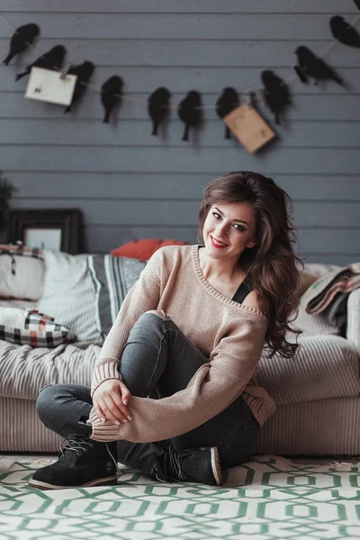 Schöne junge Frau posiert in braunem Strickpullover auf dem Teppichboden. graue Wand mit Vogelgirlanden auf Hintergrund — Stockfoto