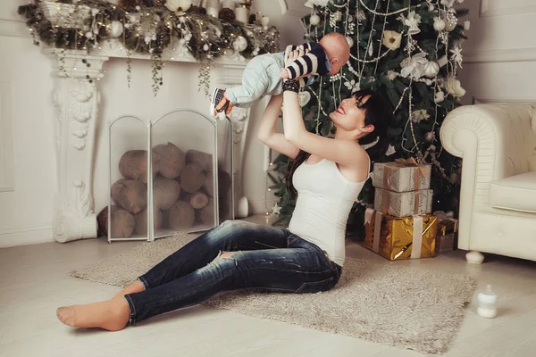 Retrato de jovem mãe feliz com criança brincando perto da árvore de Natal e lareira. Mulher segura bebê acima da cabeça — Fotografia de Stock