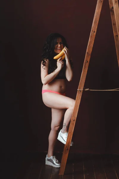 Mulher jovem grávida em forma olhando, segurando bananas na escada no estúdio loft — Fotografia de Stock