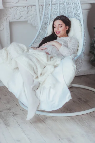 Beautiful pregnant woman sitting on wicker cocoon chair at home